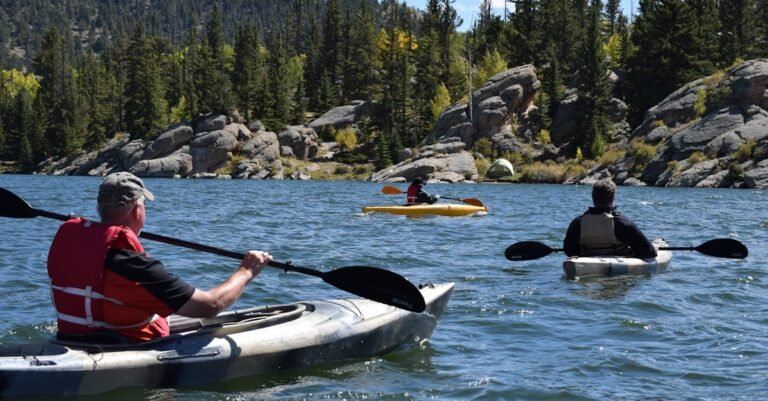 Three people kayaking on a picturesque lake surrounded by rocky terrain and evergreen trees.